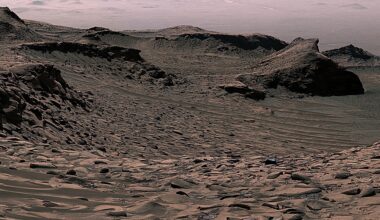 An image of Mars shows rocks and small sand dunes, with a larger mountain range in the background. Two wheel tracks in the foreground show Curiosity rover's path before it captured the image.