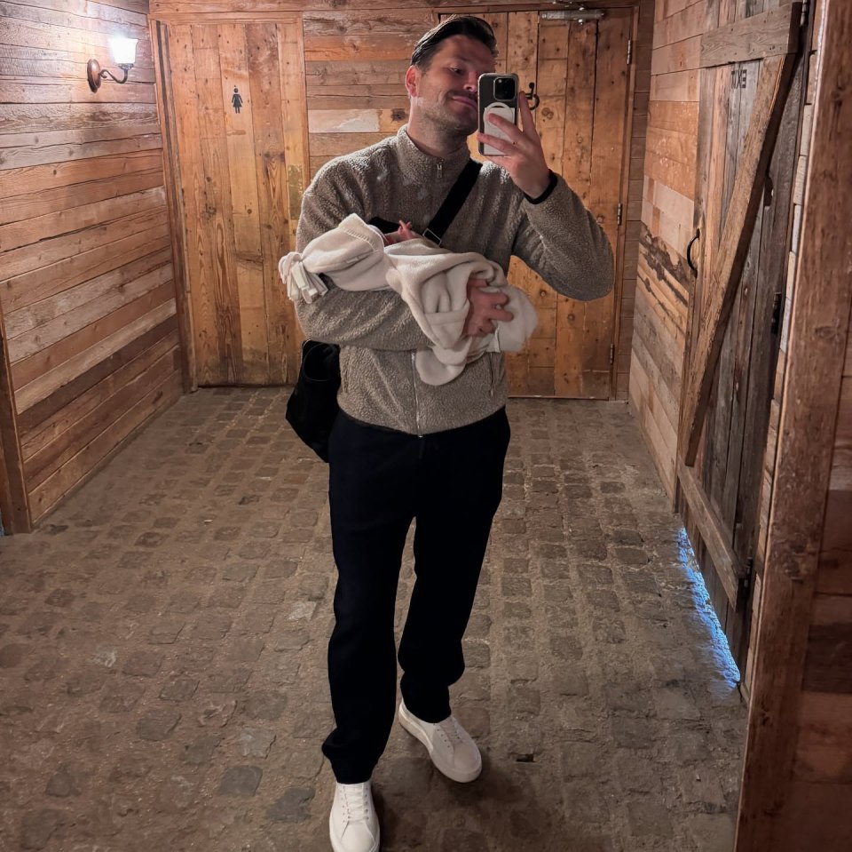 Man holding a baby and taking a selfie in a rustic hallway.