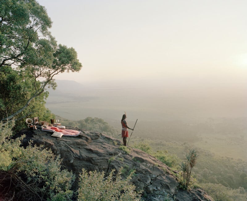A person in traditional attire stands on a rocky cliff overlooking a vast, misty valley. Nearby, blankets and cushions are arranged on the ground, suggesting a scenic resting spot surrounded by lush greenery.