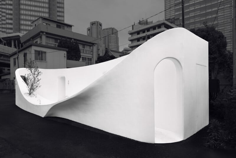 A modern, sculptural public restroom with smooth, curved white walls and arched doorways, set among urban buildings and high-rises in a cityscape.