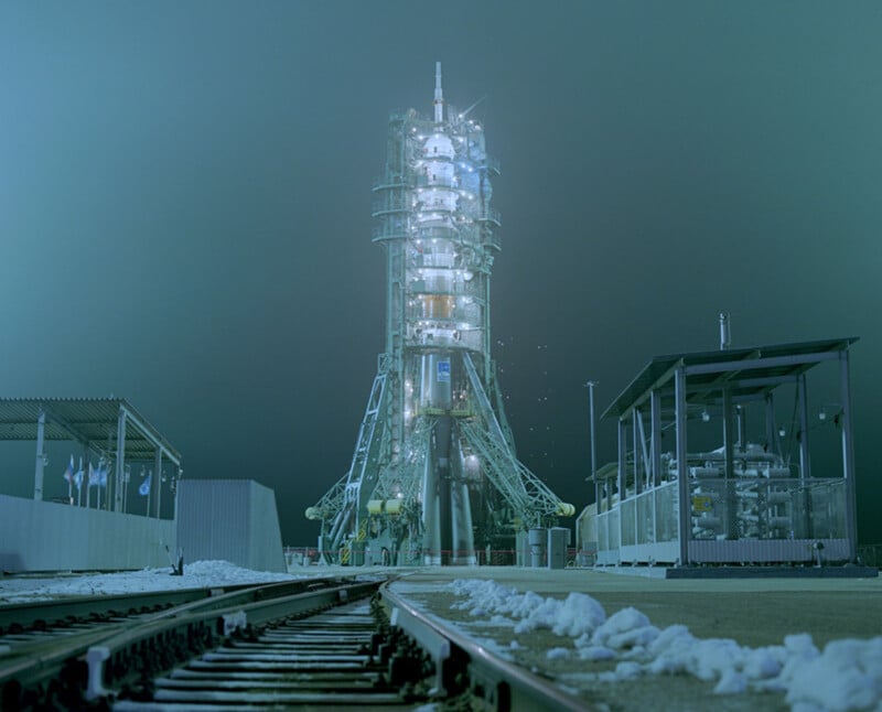 A rocket launch pad at night, illuminated by bright lights, stands surrounded by snow with train tracks leading toward it; the scene has a cold, teal-colored atmosphere.