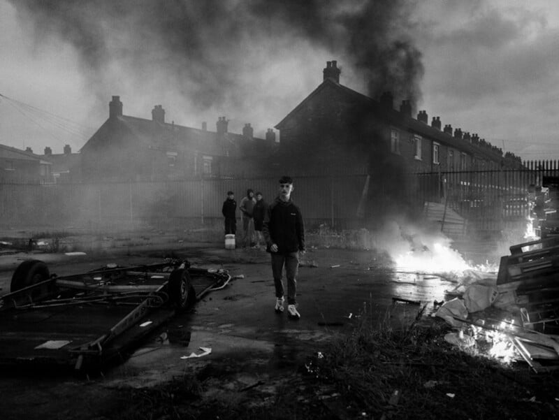 A young person walks through a desolate urban area with burning debris and thick smoke in the background, while several others stand near row houses under a cloudy sky. The scene appears tense and chaotic.
