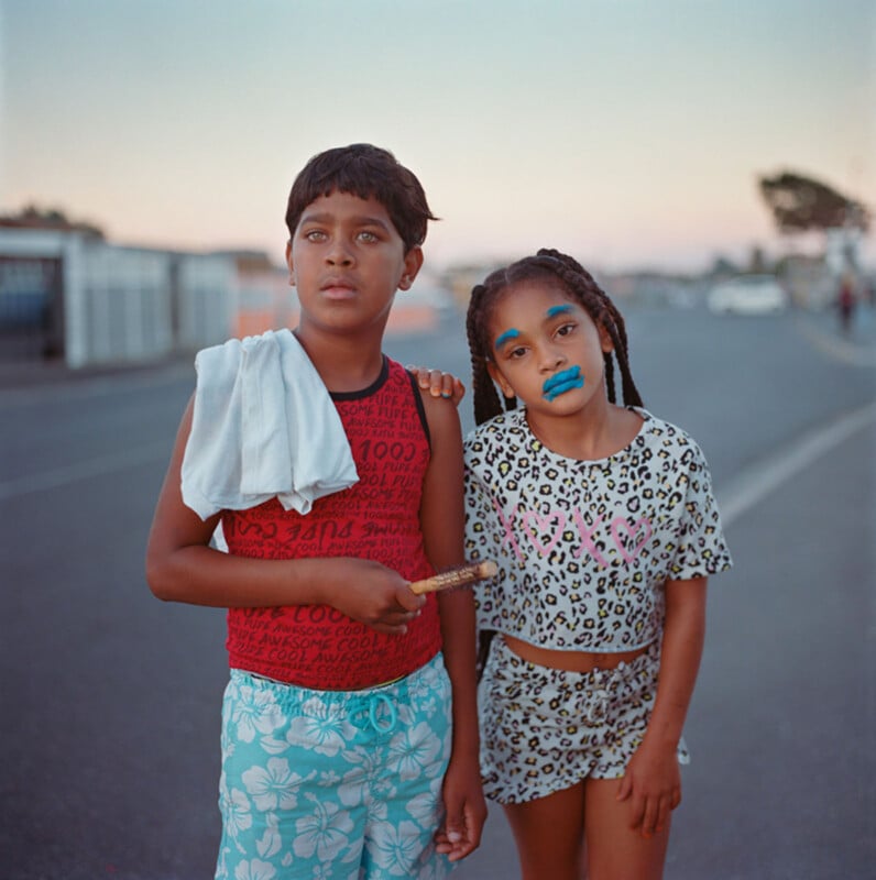 Two children stand side by side on a street at dusk. The boy wears a red shirt, blue floral shorts, and has a white cloth over his shoulder. The girl wears a leopard print outfit with blue lipstick and makeup. Both look calmly at the camera.
