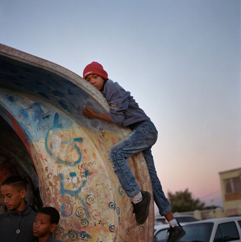 A boy in a red beanie and jeans climbs the side of a colorful, graffiti-covered concrete structure while two other boys stand nearby. Cars and buildings are visible in the background at dusk.