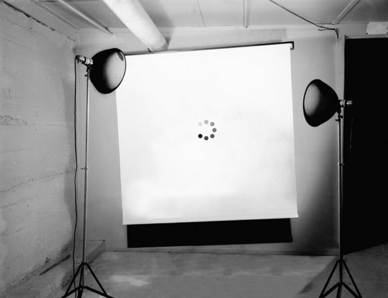 A white projection screen with a loading symbol is set up in a bare room, flanked by two studio lights on stands. The scene is minimalist and lit in black and white.