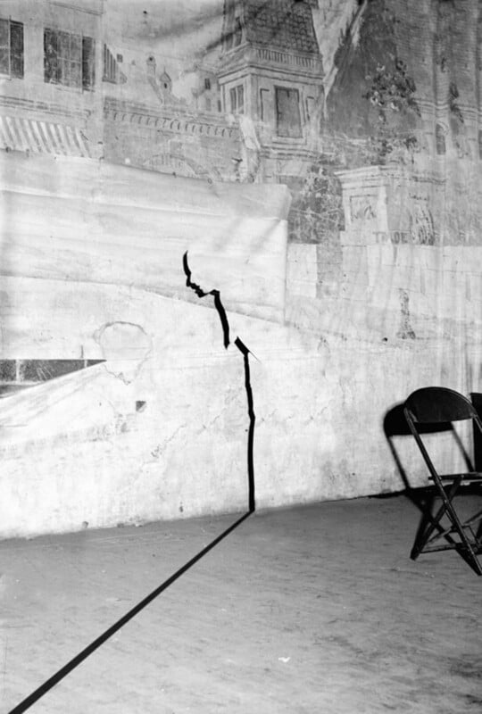 A black crack runs from the floor up a damaged wall with a faded mural. Two empty folding chairs sit on the right side, casting shadows on the floor.