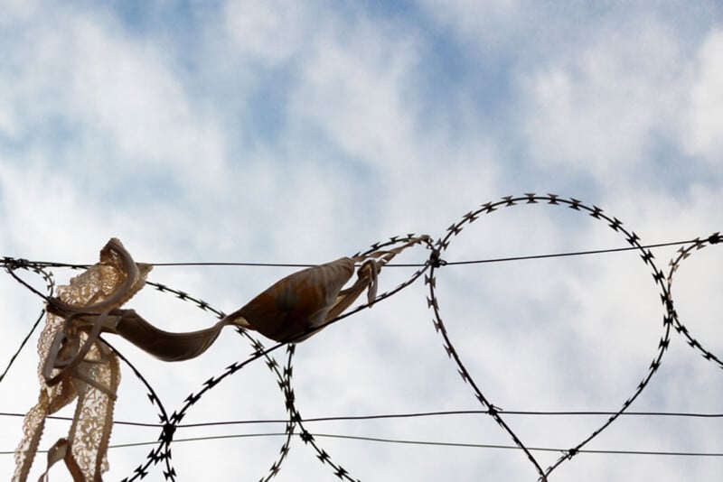 A bra and a piece of lace are caught on the coils of a barbed wire fence under a cloudy sky.