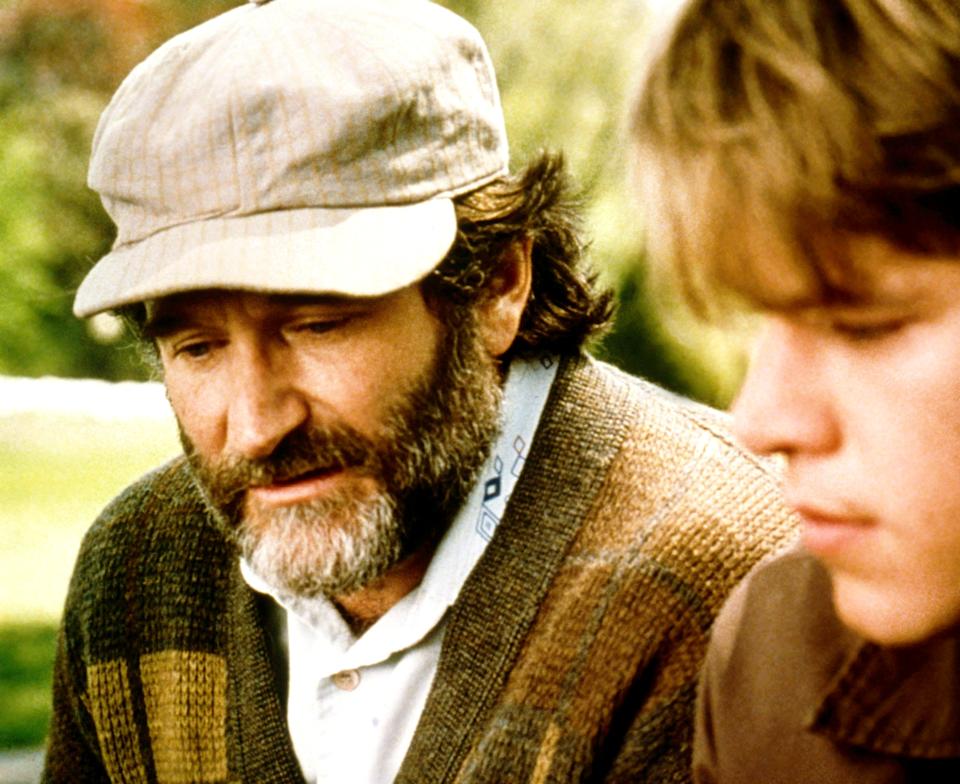 Two men sit closely, one older with a beard and cap, the other younger with short hair, appearing thoughtful and engaged in conversation