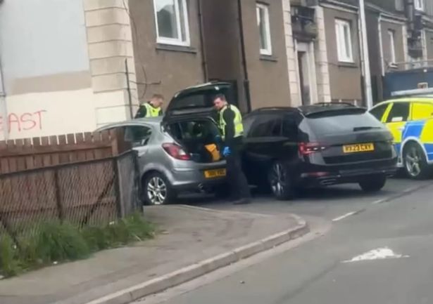 An image of uniformed police officers searching the boot of a grey car.