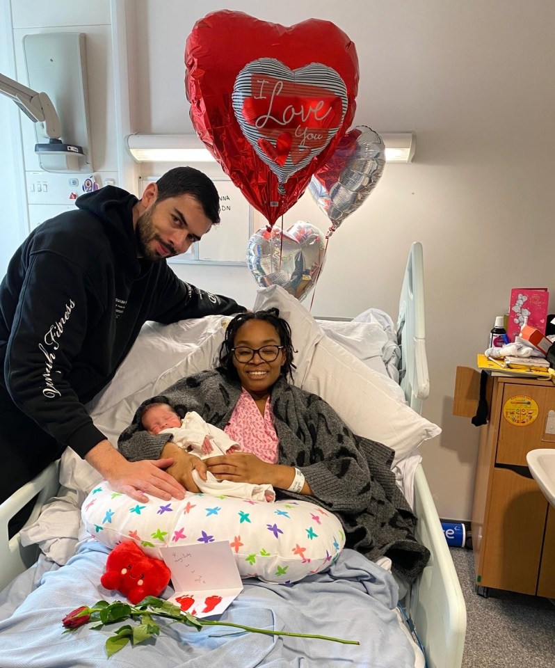 A new mother and father in a hospital room with their newborn baby.  Heart-shaped balloons are in the background.