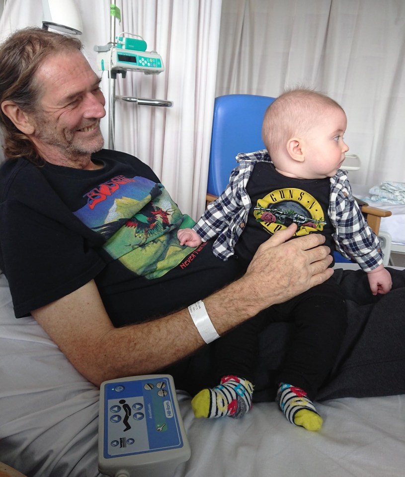 A father in a hospital bed holds his baby, who is wearing a Guns N' Roses t-shirt.