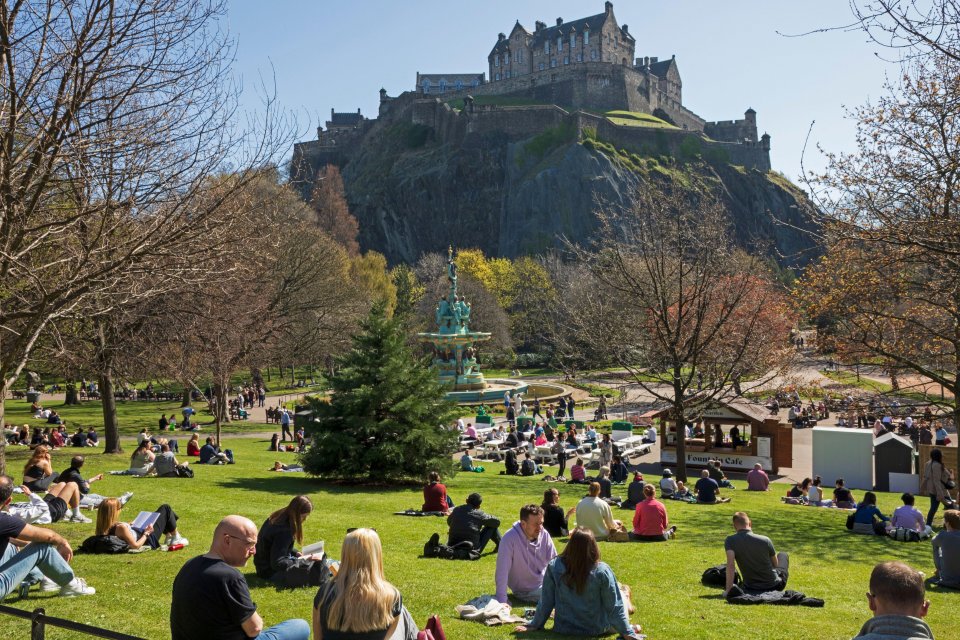 People enjoying a sunny day in Princes Street Gardens in Edinburgh, Scotland, with Edinburgh Castle in the background.