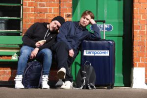 Comedian Joe Thomas and another man sitting at a train station, appearing bored.