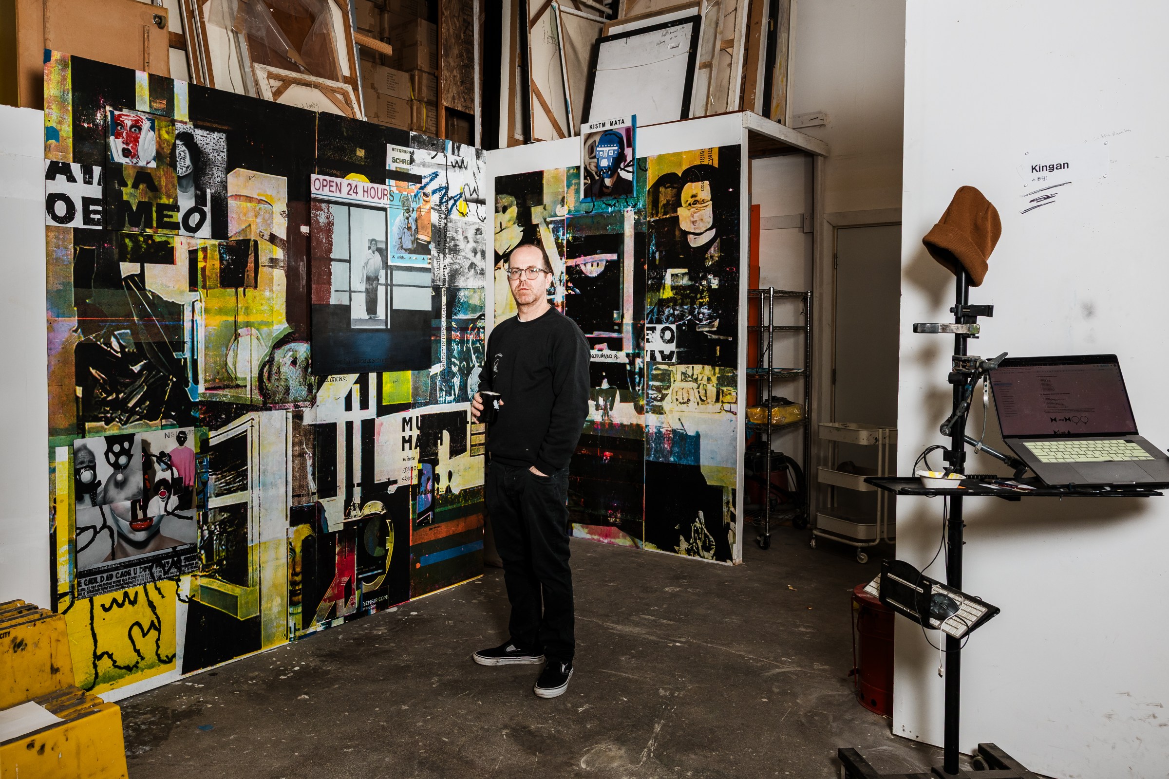 A man stands in the center of the frame in an art studio filled with his paintings.