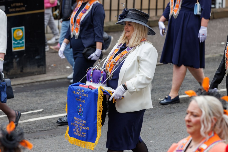 Woman in a white jacket carrying a crown and a blue banner at an Orange Order parade.