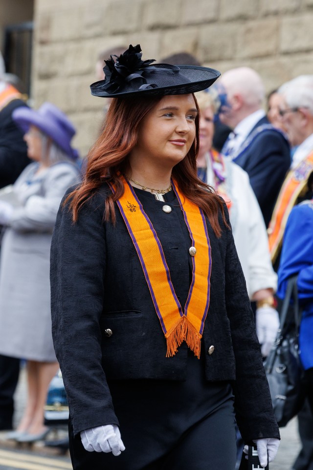 Woman in black jacket and hat wearing an orange sash.