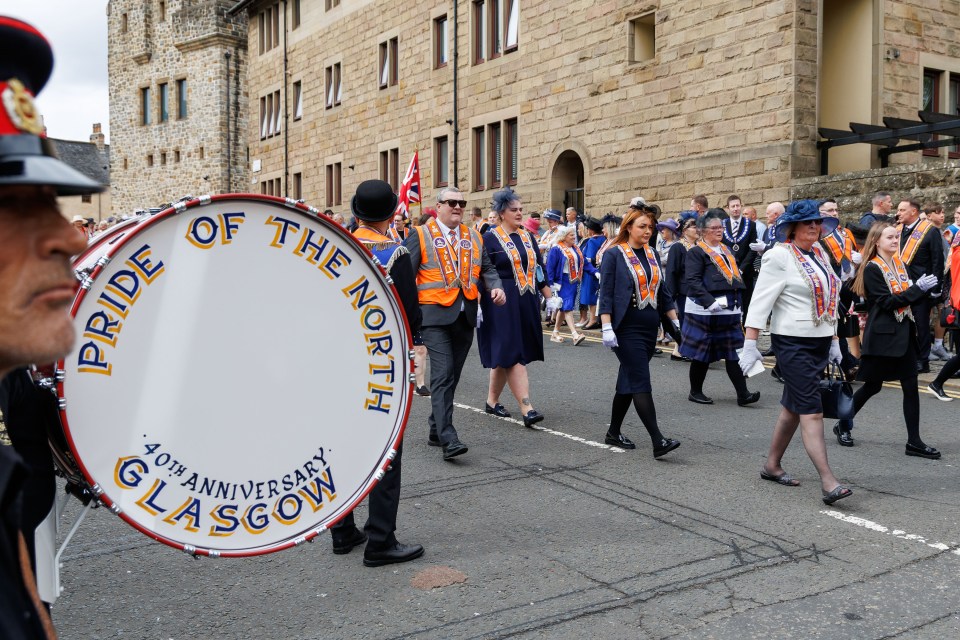 A parade featuring people wearing orange sashes and a bass drum that reads "Pride of the North, 40th Anniversary, Glasgow".