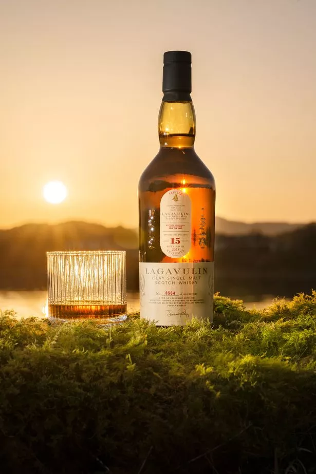A bottle of Lagavulin’s special edition 15 Year Old whisky beside a glass outside with a setting sun in the distance
