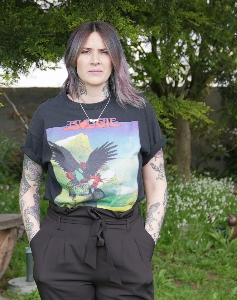 Woman with tattoos wearing a black t-shirt and black pants, standing outdoors.