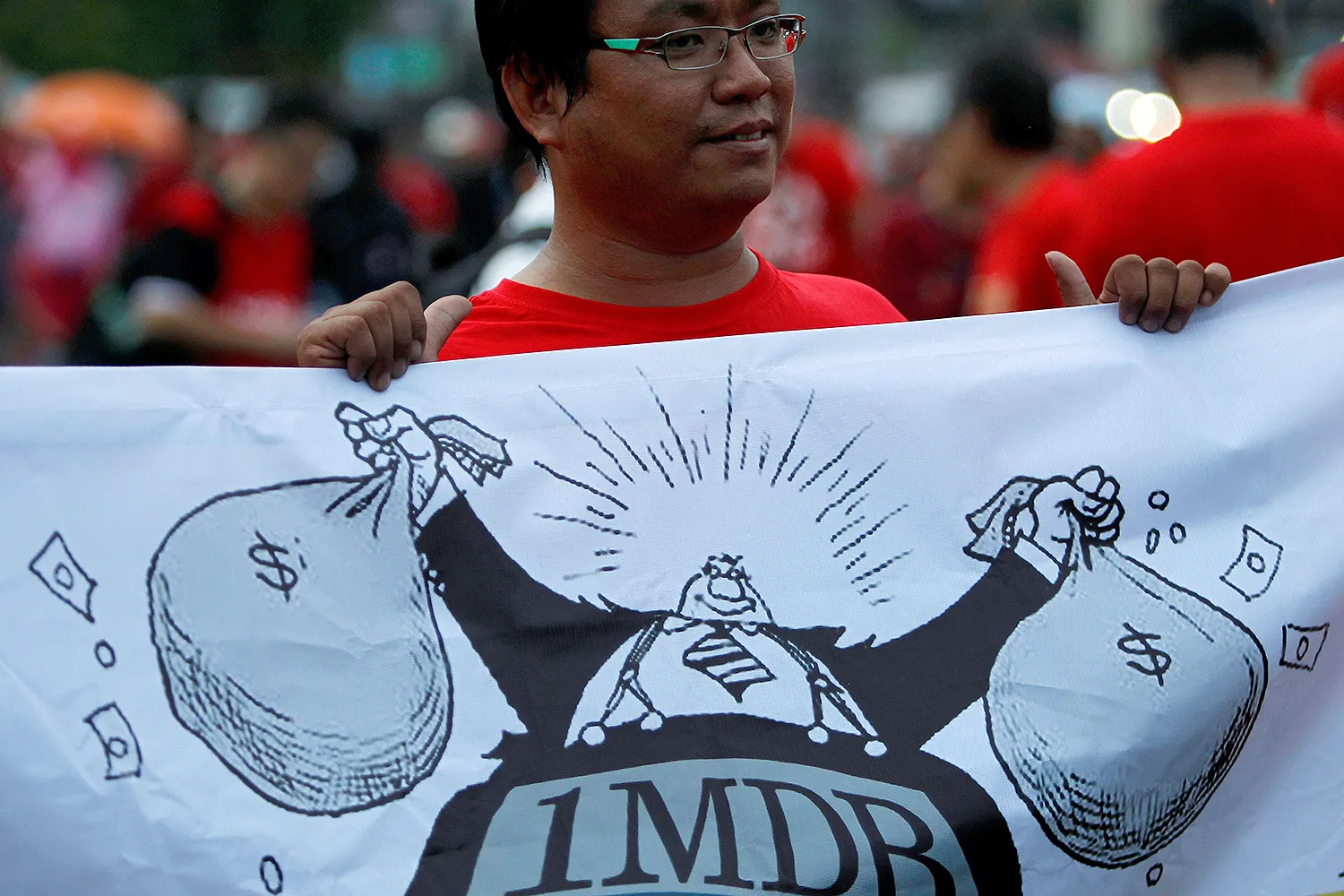 A man holds up a banner of a cartoon man holding bags of money with "1MBD" on his clothes.