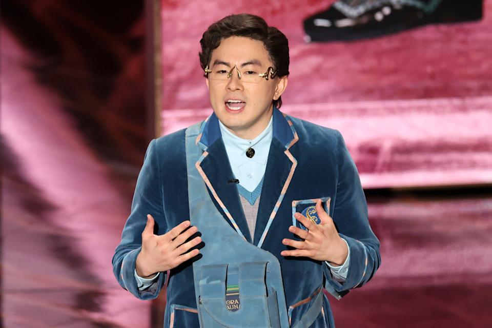 Bowen Yang in a velvet suit speaks animatedly on stage, gesturing with hands