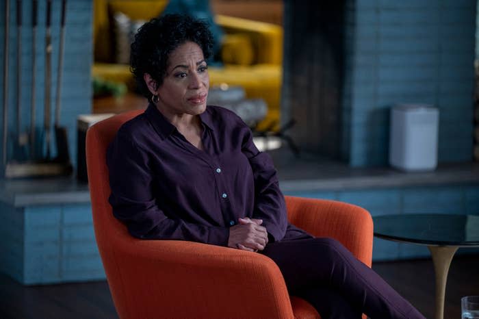 Person seated in an orange chair, wearing a dark, formal outfit, looking thoughtful in a modern indoor setting