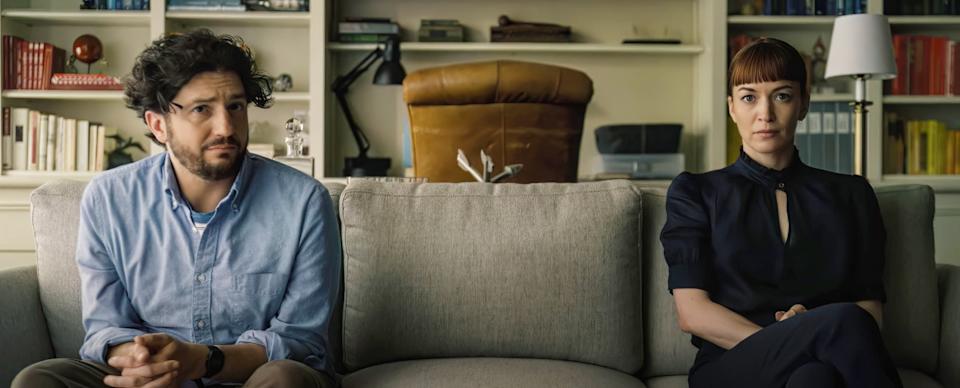 A man and woman sit on a sofa with neutral expressions, in a room with bookshelves in the background