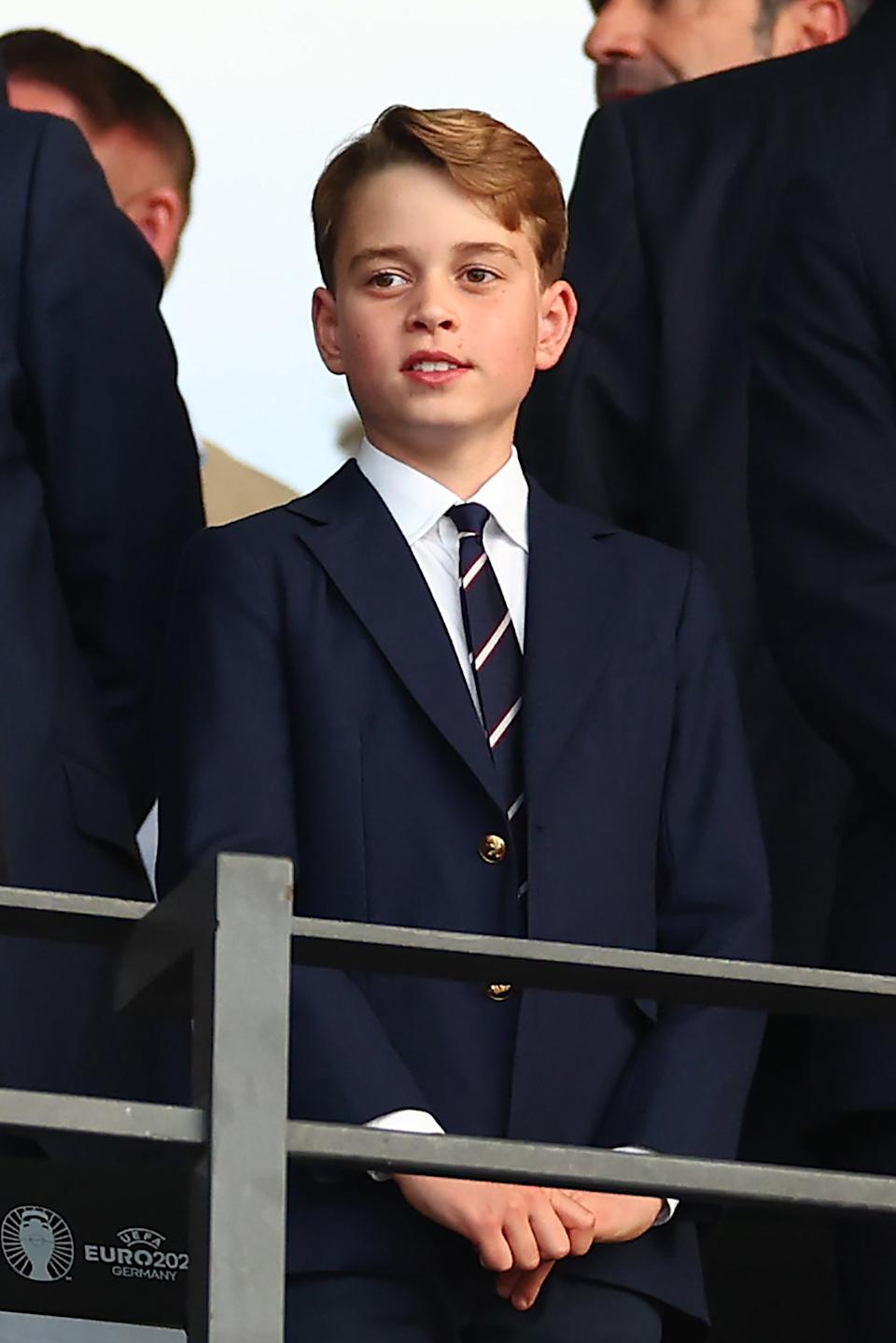 Prince George in a blue suit and striped tie looking up from a crowd.
