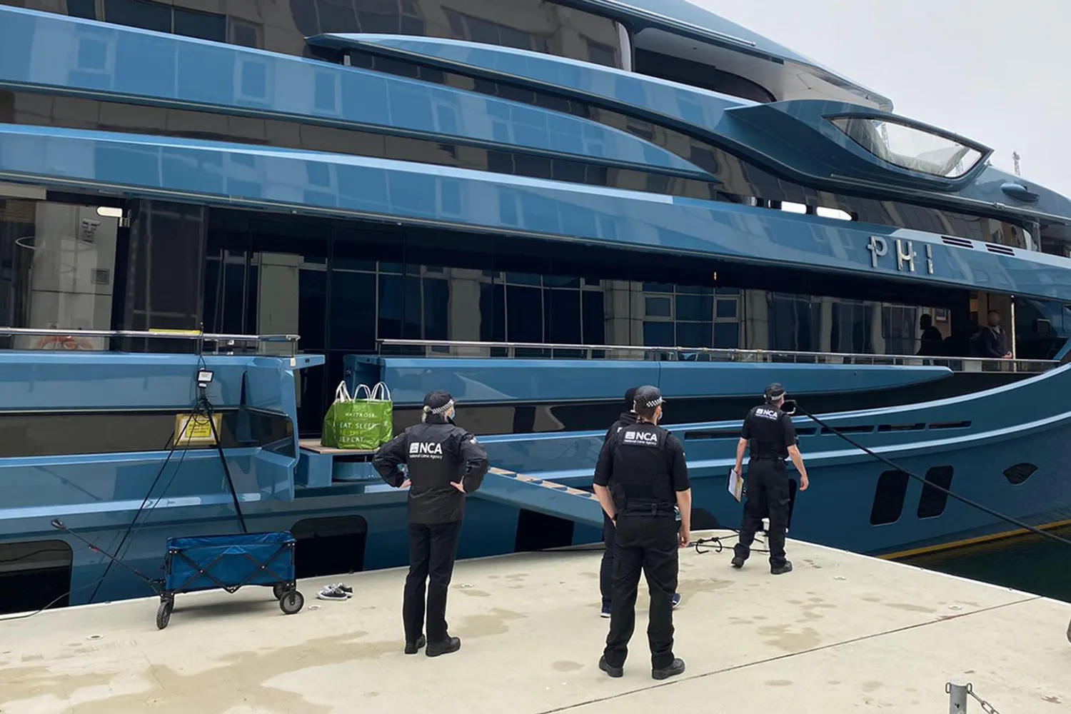 Members of London's National Crime Agency stand before a massive yacht called "Phi" at a dock.
