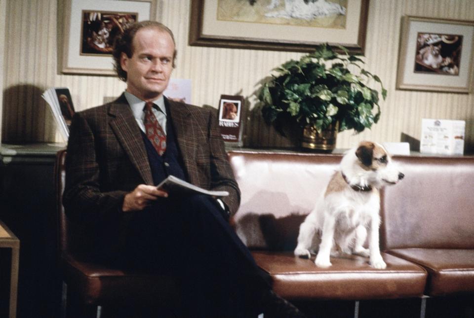A man in a suit sits on a couch with a small dog, looking thoughtful. The setting appears to be an office with framed pictures on the wall
