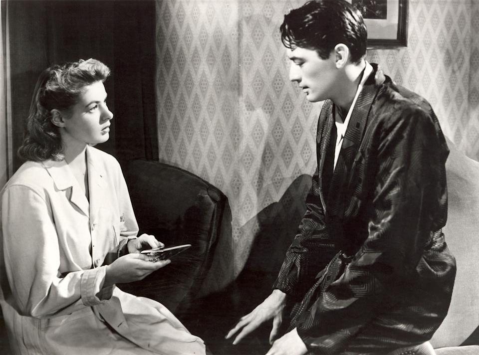 Two people in a classic film scene, one seated holding a clipboard, while the other, wearing a robe, sits attentively