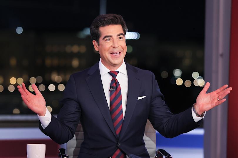 Jesse Watters in a suit and striped tie gestures with open arms, sitting in a studio with blurred lights in the background