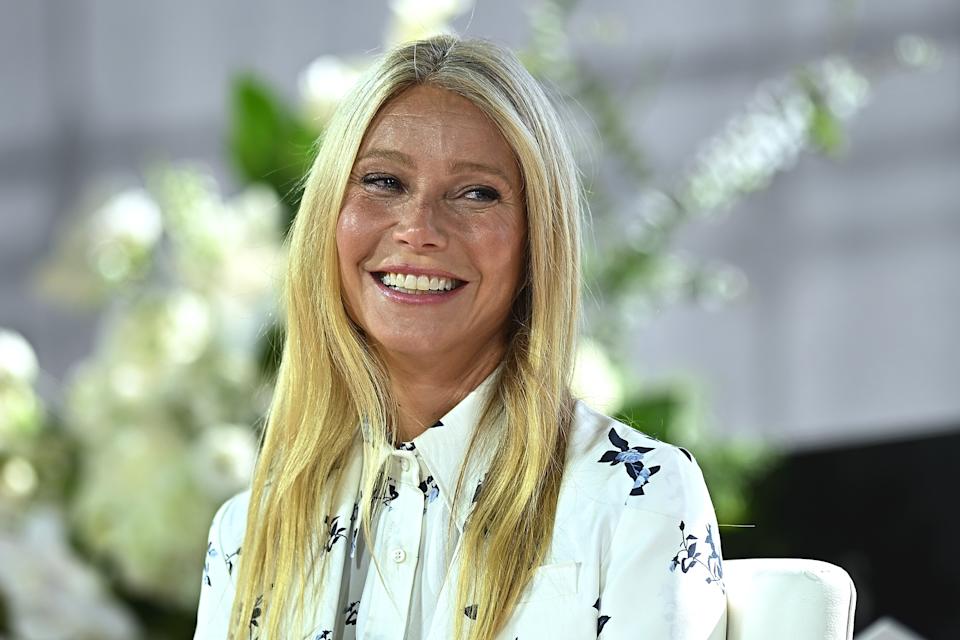 Person smiling, wearing a floral-patterned blouse, in a well-lit, elegant setting with floral decorations in the background