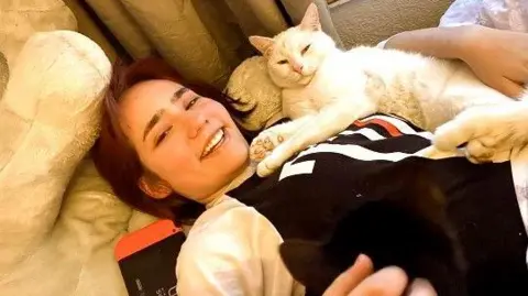 Family handout Elise Sebastian lies on a sofa with her cat on her. It is a white short haired cat. Elise is smiling and wearing a long sleeved black and white top.