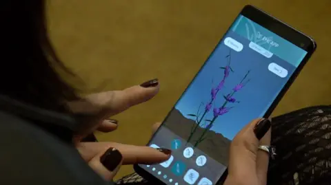 A mobile phone is being held and on the screen is an image of a digitally created purple plant on a blue background. The person holding the phone has black nail polish on.