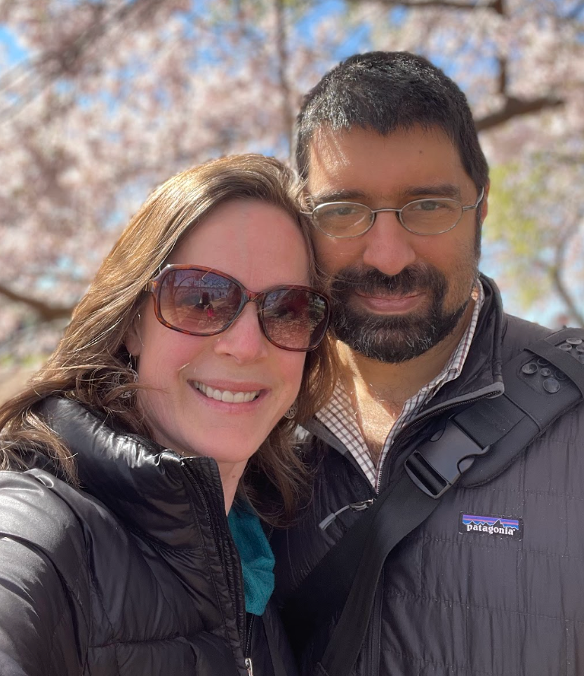 Two people in sunglasses and jackets smiling outdoors in front of blossoming trees