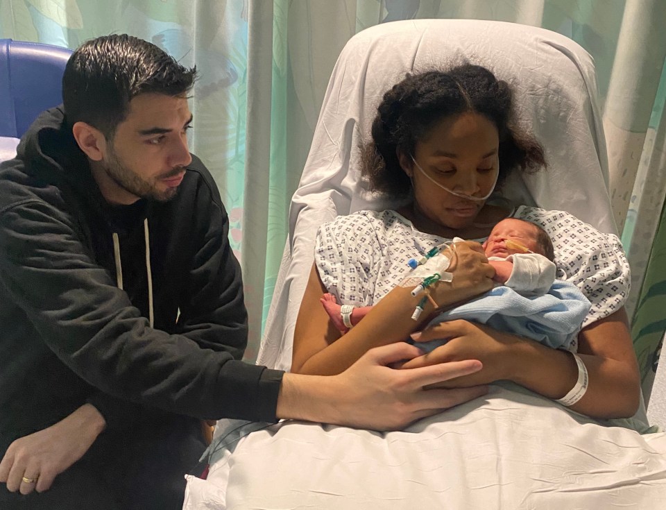 A mother holding her newborn baby in a hospital bed, with her husband sitting beside them.