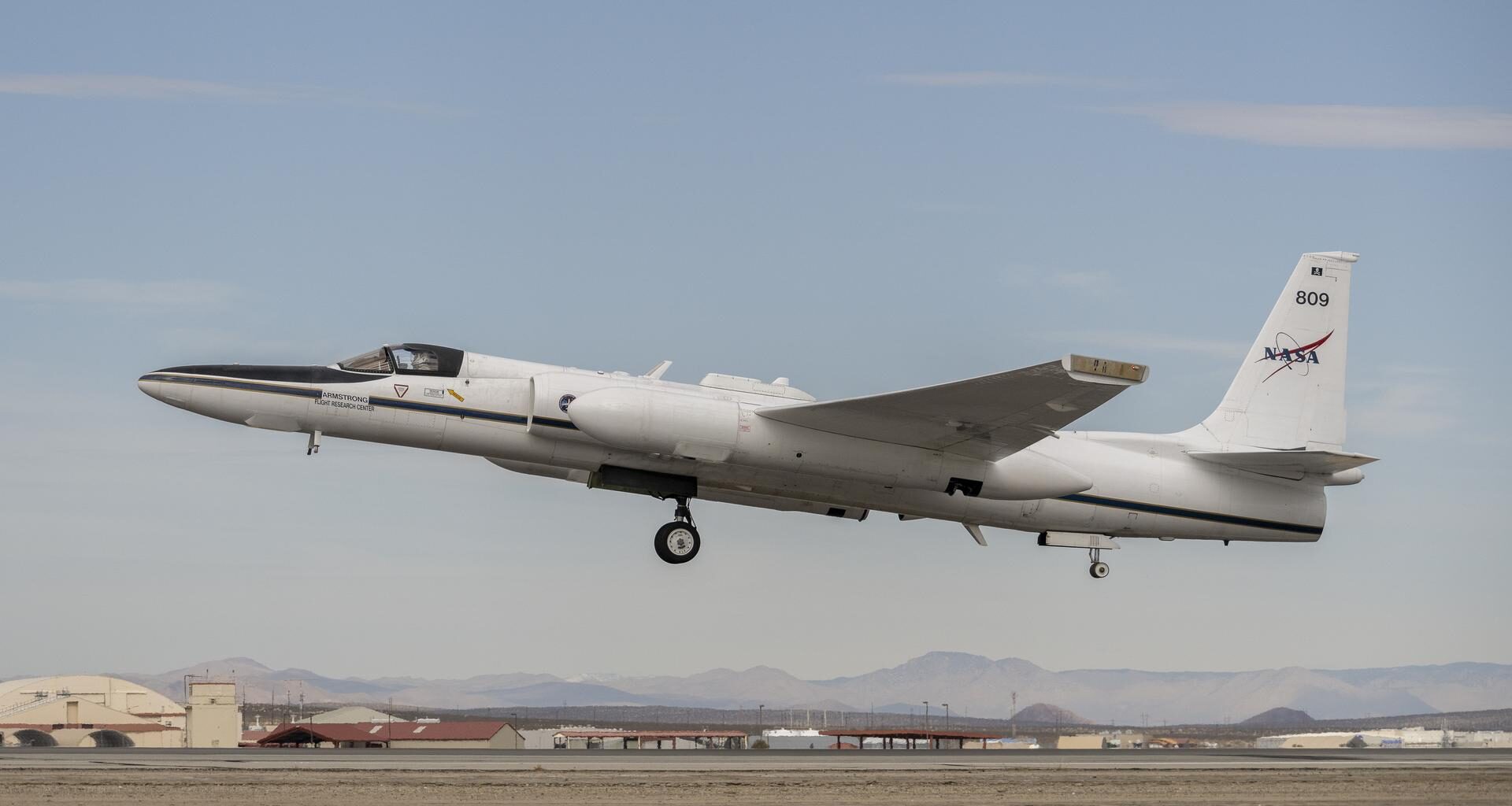 A plane begins to ascend from the runway on dim desert morning, heading toward the left of the frame. The wheels of the plane are still dropped and the nose of the plane is pointed upward toward higher skies, its tail featuring the NASA insignia. The helmeted pilot inside is barely visible through the cockpit window. Below a desert landscape with indiscernible buildings are flanked by a hazy mountain range in the distance.