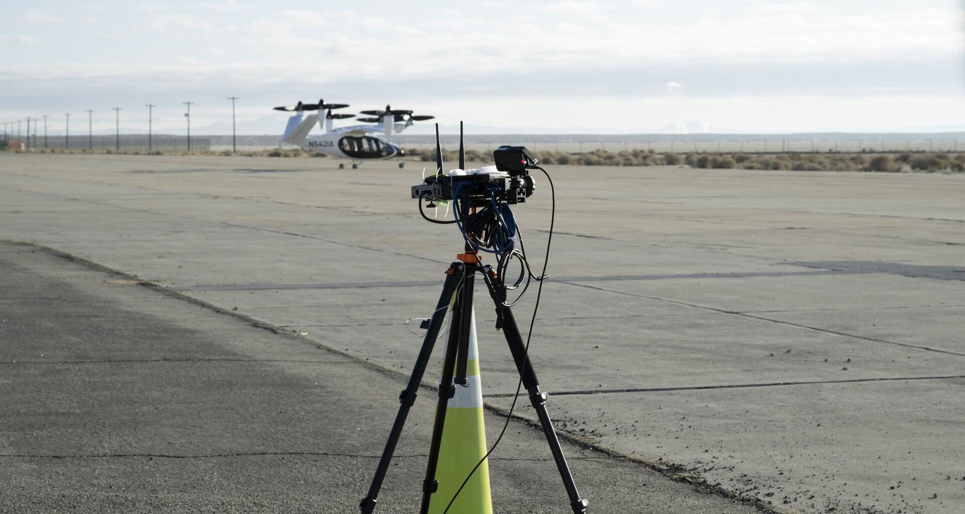 A yellow traffic cone and a black tripod with black testing instruments stands in the foreground on a concrete pad with a desert landscape, power lines, and a black and white aircraft in the background. The aircraft has six black propellors that sit on white arms and connect to the aircraft body, which has black doors and is pod-shaped. The aircraft sits on three small wheels.