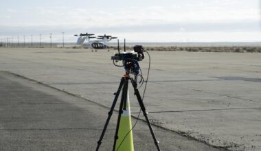A yellow traffic cone and a black tripod with black testing instruments stands in the foreground on a concrete pad with a desert landscape, power lines, and a black and white aircraft in the background. The aircraft has six black propellors that sit on white arms and connect to the aircraft body, which has black doors and is pod-shaped. The aircraft sits on three small wheels.