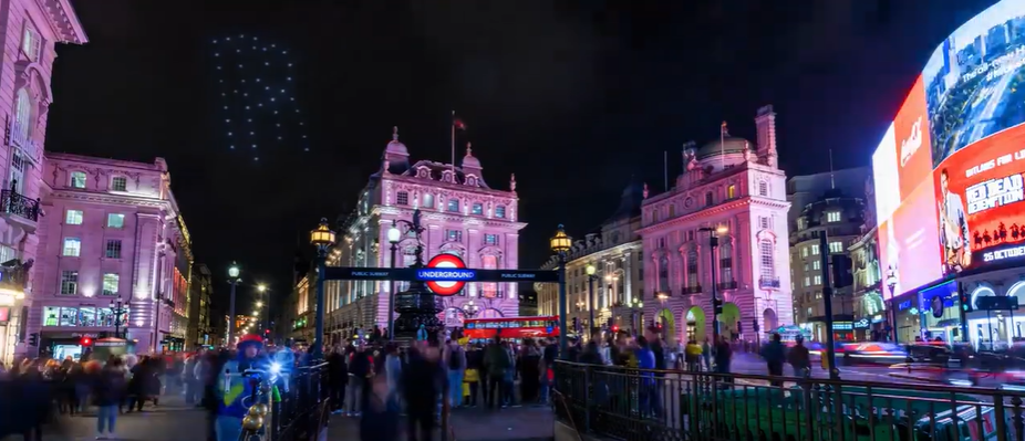 Nighttime view of Piccadilly Circus in London with drone lights in the sky.