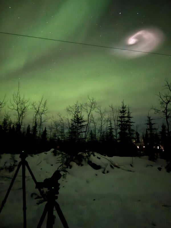 A snowy landscape with silhouetted trees under the green glow of the Northern Lights. A bright spiral pattern appears in the sky. A camera on a tripod is in the foreground, capturing the scene.