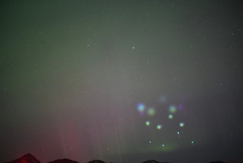 A night sky featuring the aurora borealis with green and red hues, stars scattered throughout. Faint, colorful lights are visible near the center of the image above the silhouetted horizon of rolling hills.