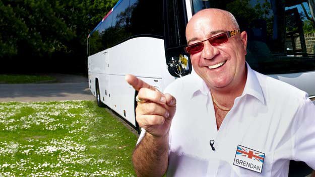 Coach Trip with Brendan Sheerin. He is stood in front of a white coach and he is pointing while smiling.