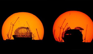Two large telescope domes under construction are silhouetted against a bright orange sunset, with several cranes visible around the structures.