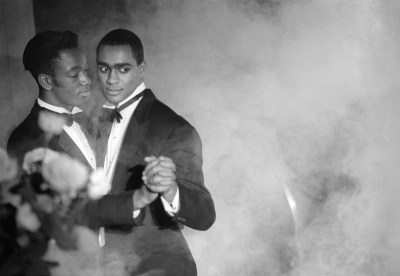 Two Black men in a suits dancing in a smoky room.