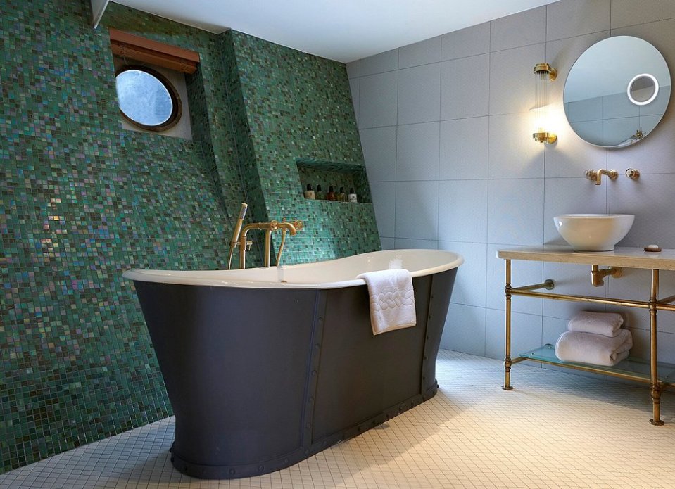 Bathroom with dark freestanding tub, green mosaic tile wall, and brass fixtures.