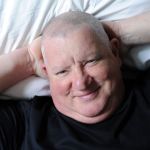 A man lies back on a bed with his hands behind his head.