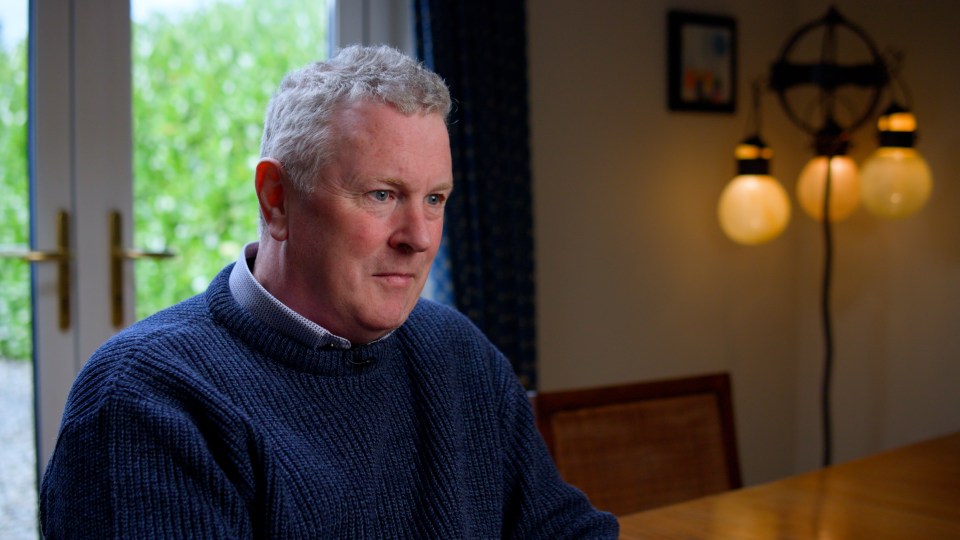 A man in a blue sweater sits and looks thoughtfully to his right.