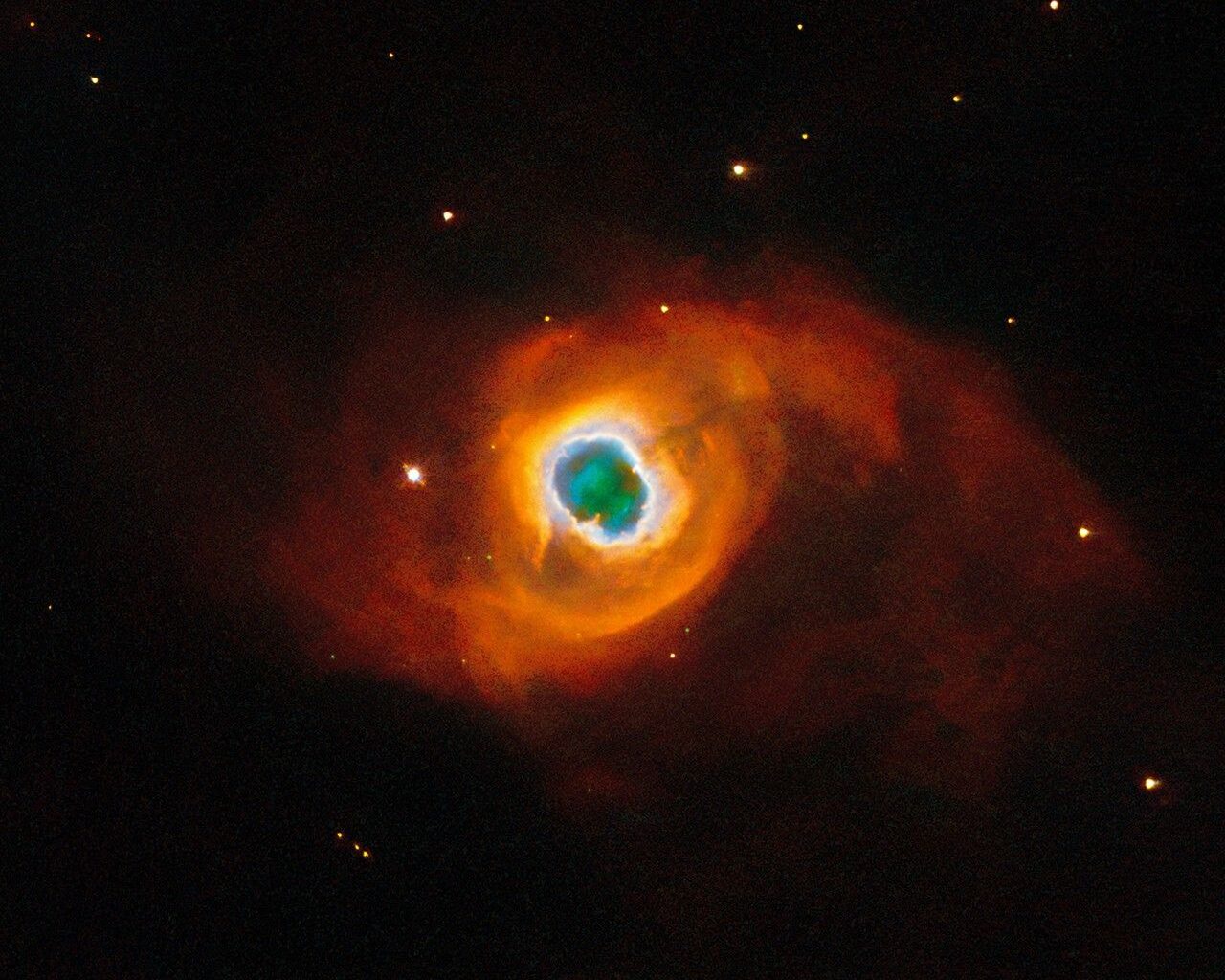 The image holds a planetary nebula, a glowing shell of material thrown off by a dying star. A glowing, jagged ring encircles a small central region of greenish clouds, making it appear like a hole torn in fabric. This “hole” is itself encircled by a band of silvery-blue clouds and a larger, fainter, yellow ring of gas. Puffy, smoky clouds of orange and red gas billow out from there into a large oval nebula, fading into the dark background of space dotted by stars.